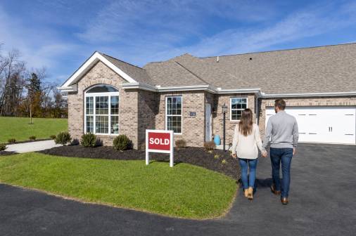A couple walks hand in hand in front of a house displaying a "Sold" sign, celebrating their new home purchase.